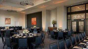 A large conference room with multiple tables set for dining, featuring black chairs and tables, a chandelier, and a large window with blinds.
