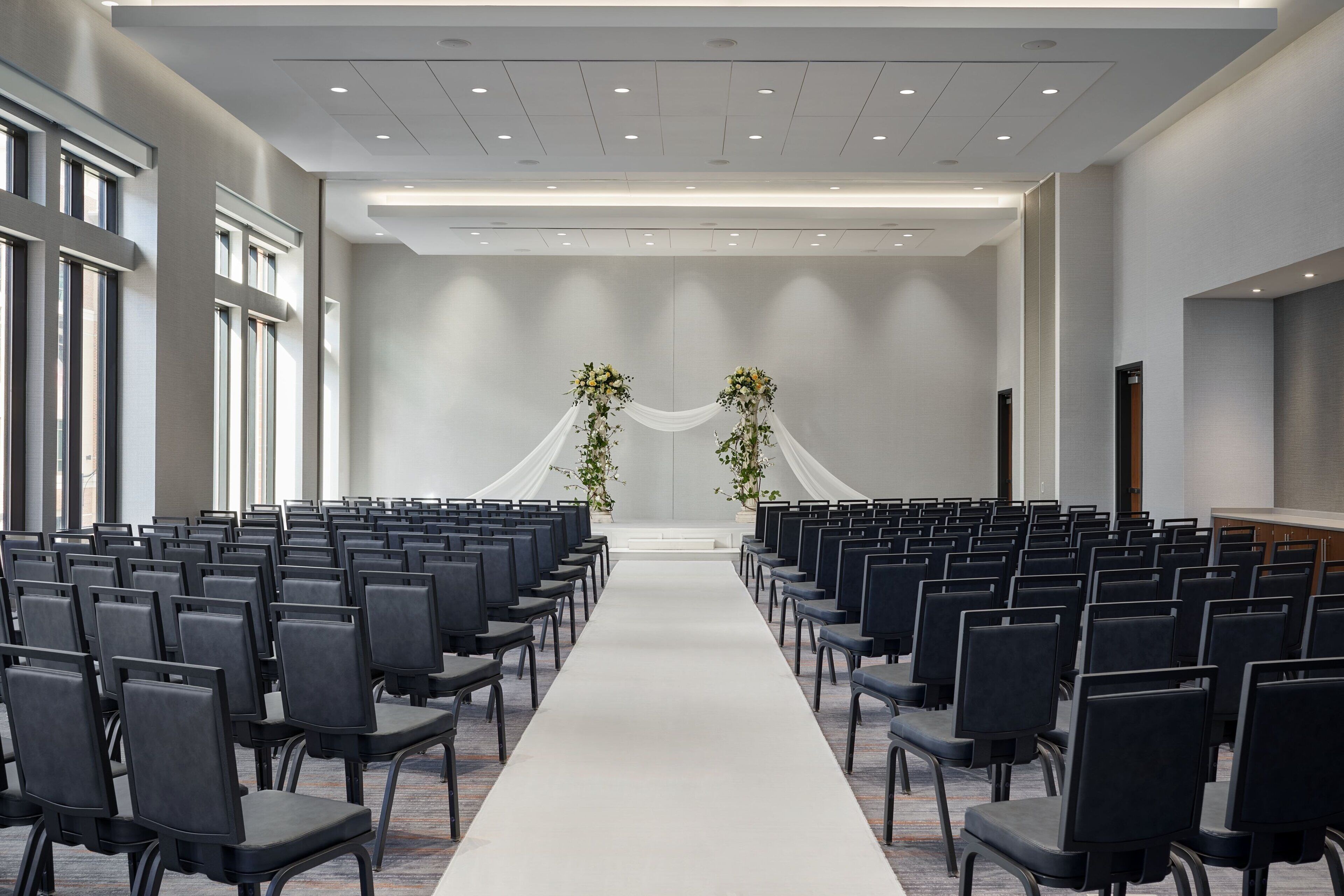 A conference room with rows of chairs, a central aisle, and a stage with draped fabric and floral arrangements.