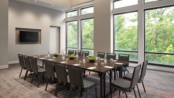 A conference room with a long table, chairs, and large windows offering a view of trees.