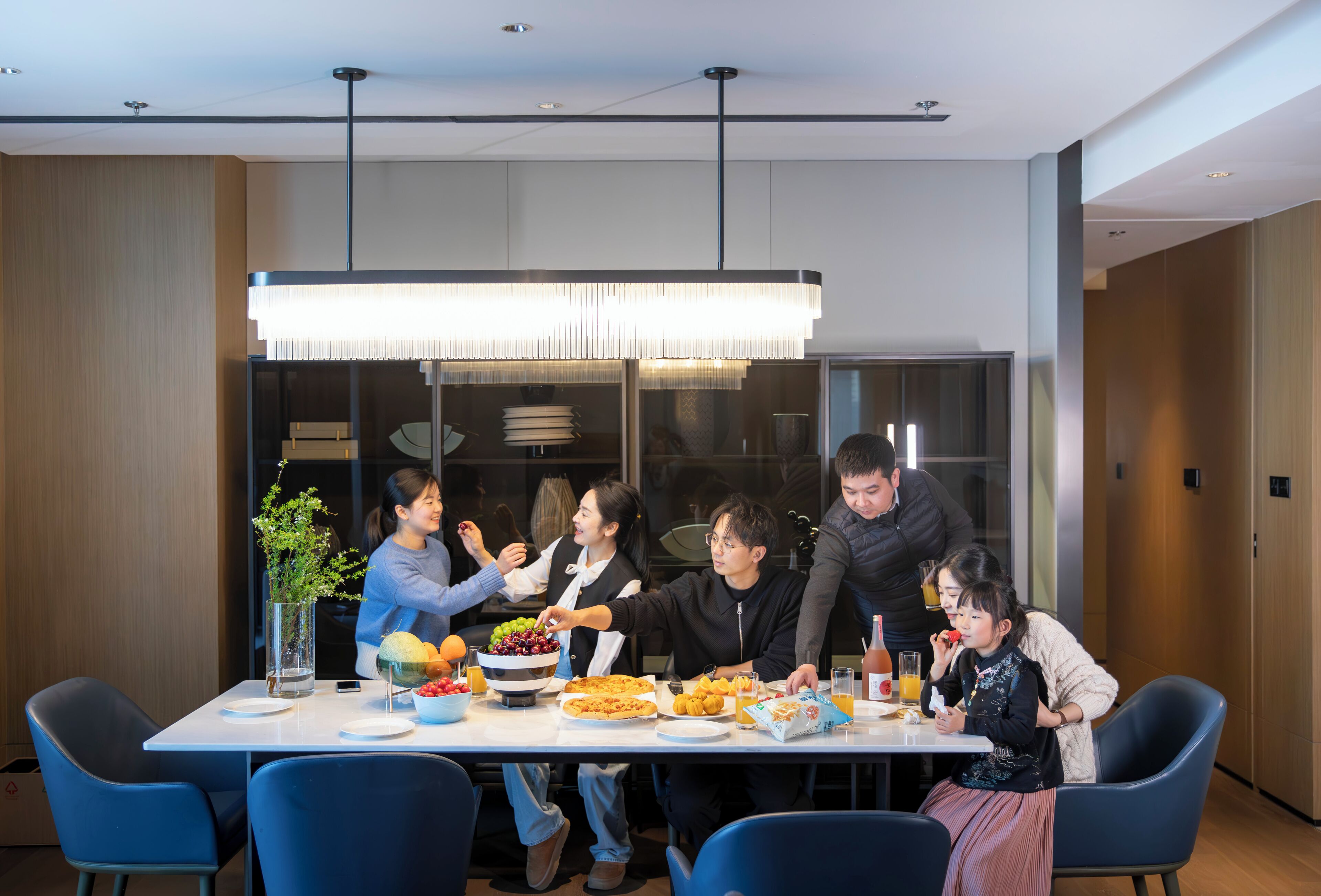 A group of people dining at a table with various dishes, a vase with a plant, and a modern pendant light.