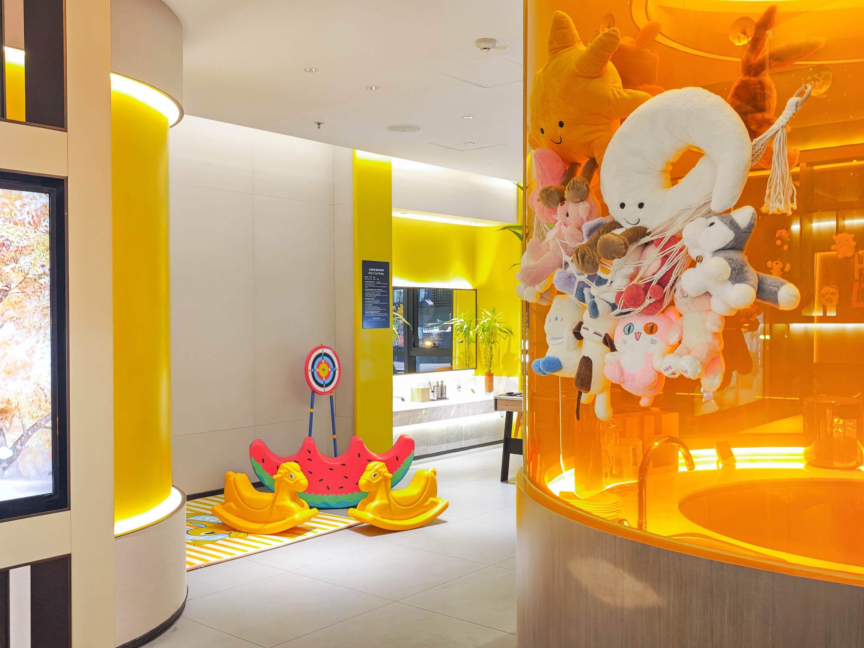 A brightly lit indoor space with a yellow and white color scheme, featuring a display of plush toys, a target board, and a children's play area with yellow rocking horses.