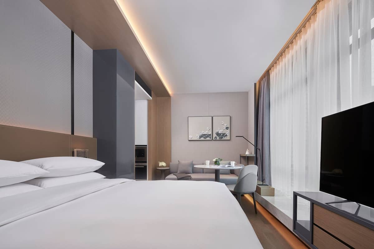 Una habitación de hotel moderna con una cama grande, televisión, zona de estar con una mesa y una ventana con cortinas.
