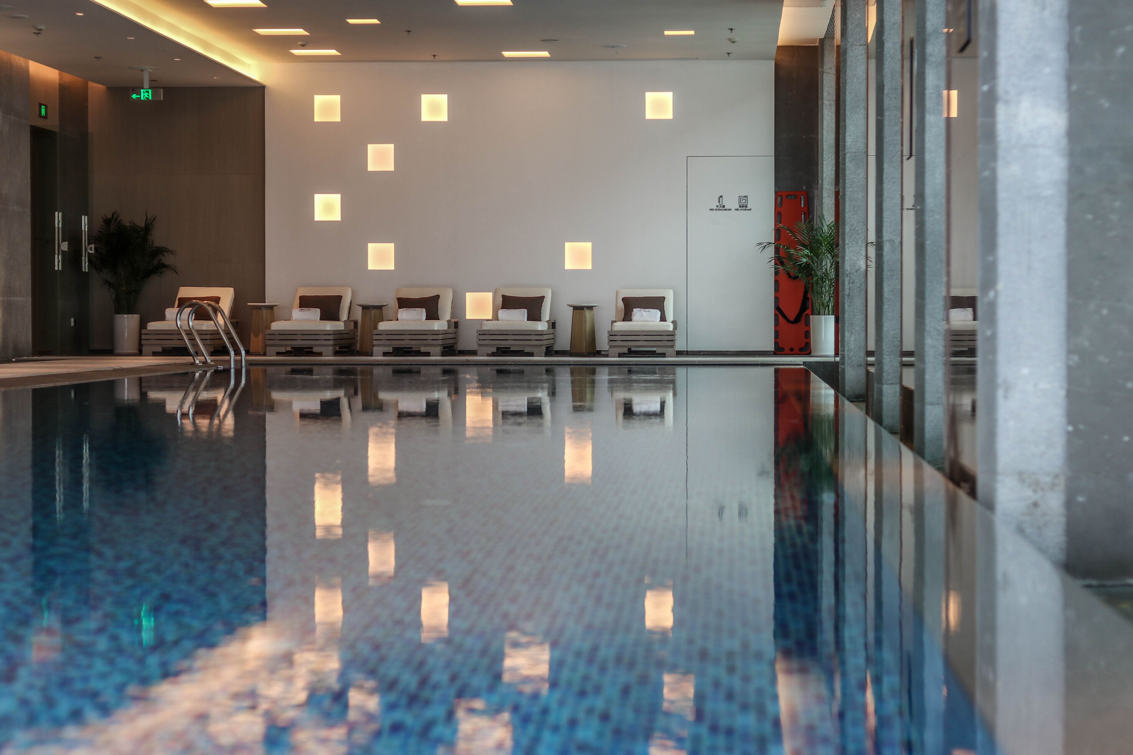 Un área de piscina interior moderna con suelo de vidrio transparente, sillas de descanso e iluminación empotrada en las paredes.