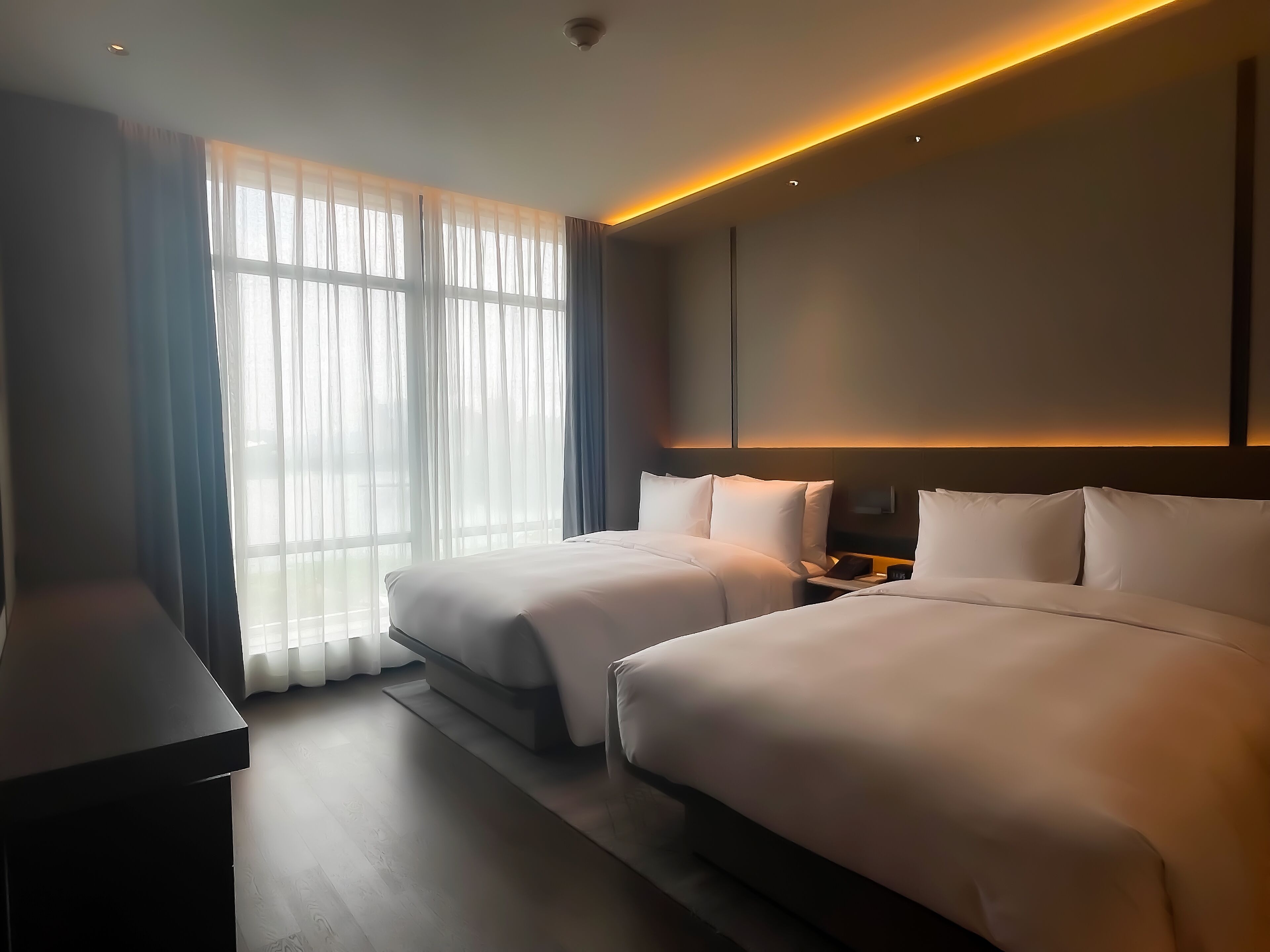 Une chambre d’hôtel moderne avec deux lits, une grande fenêtre avec des rideaux légers et un éclairage doux au plafond.