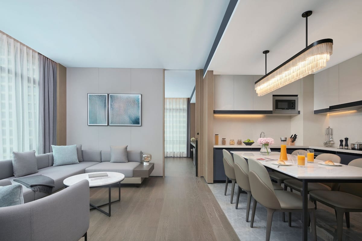 Una sala de estar moderna con sofá, mesa de centro y comedor con sillas. Se observa una cocina con microondas y una lámpara colgante.