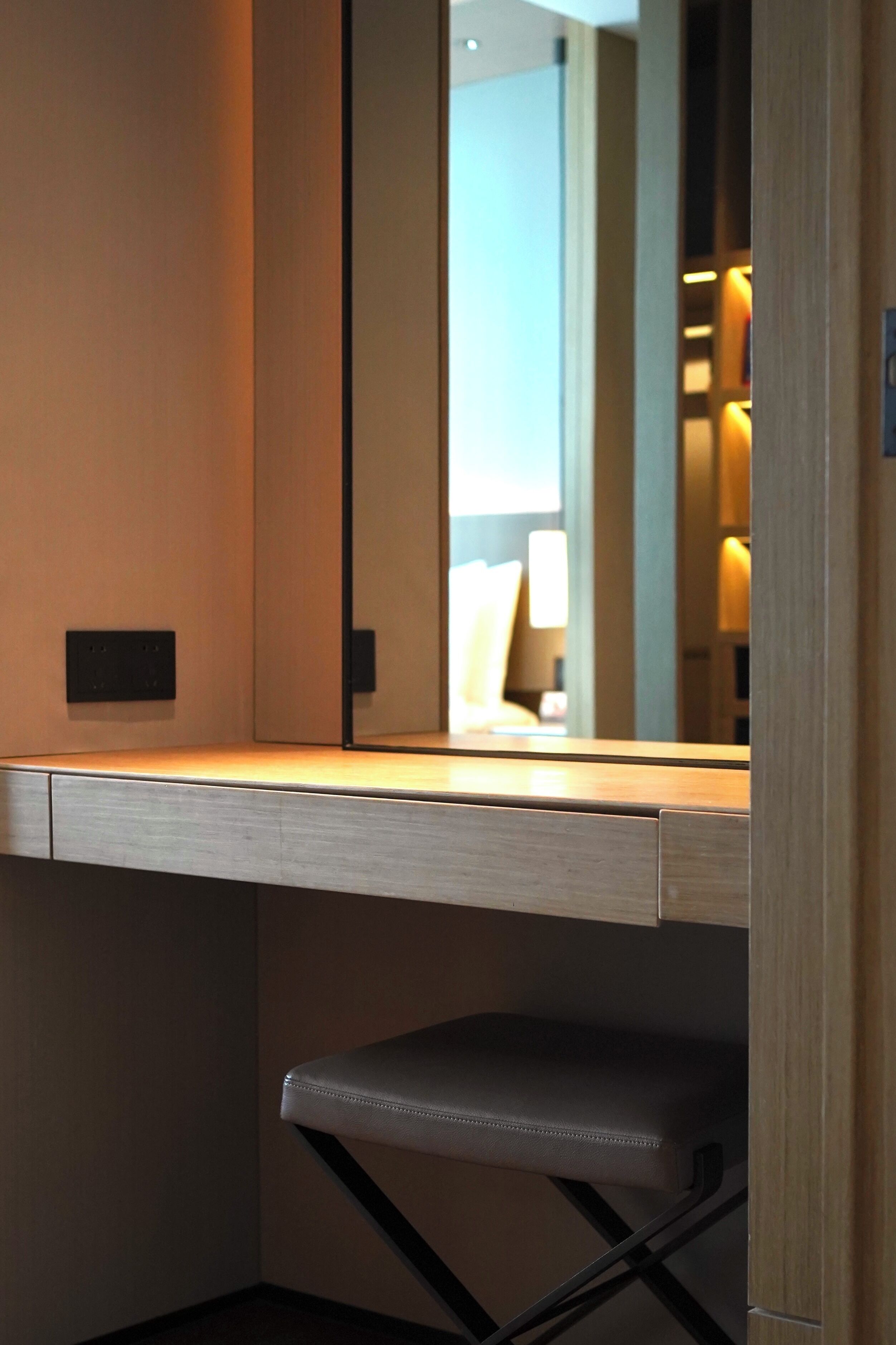 Una habitación de hotel moderna con un escritorio de madera, una silla acolchada y un espejo que refleja una habitación bien iluminada con una lámpara y una cama.