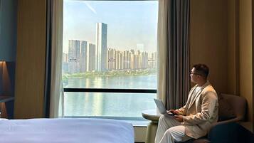 A hotel room with a large window offering a view of a city skyline and a body of water. There is a bed and a chair with a laptop on it.