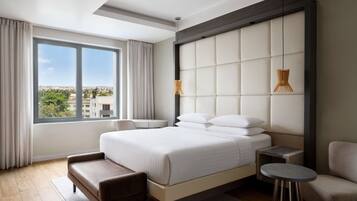 A modern hotel room with a large bed, a sitting area, a small table, and a view of the city through large windows.