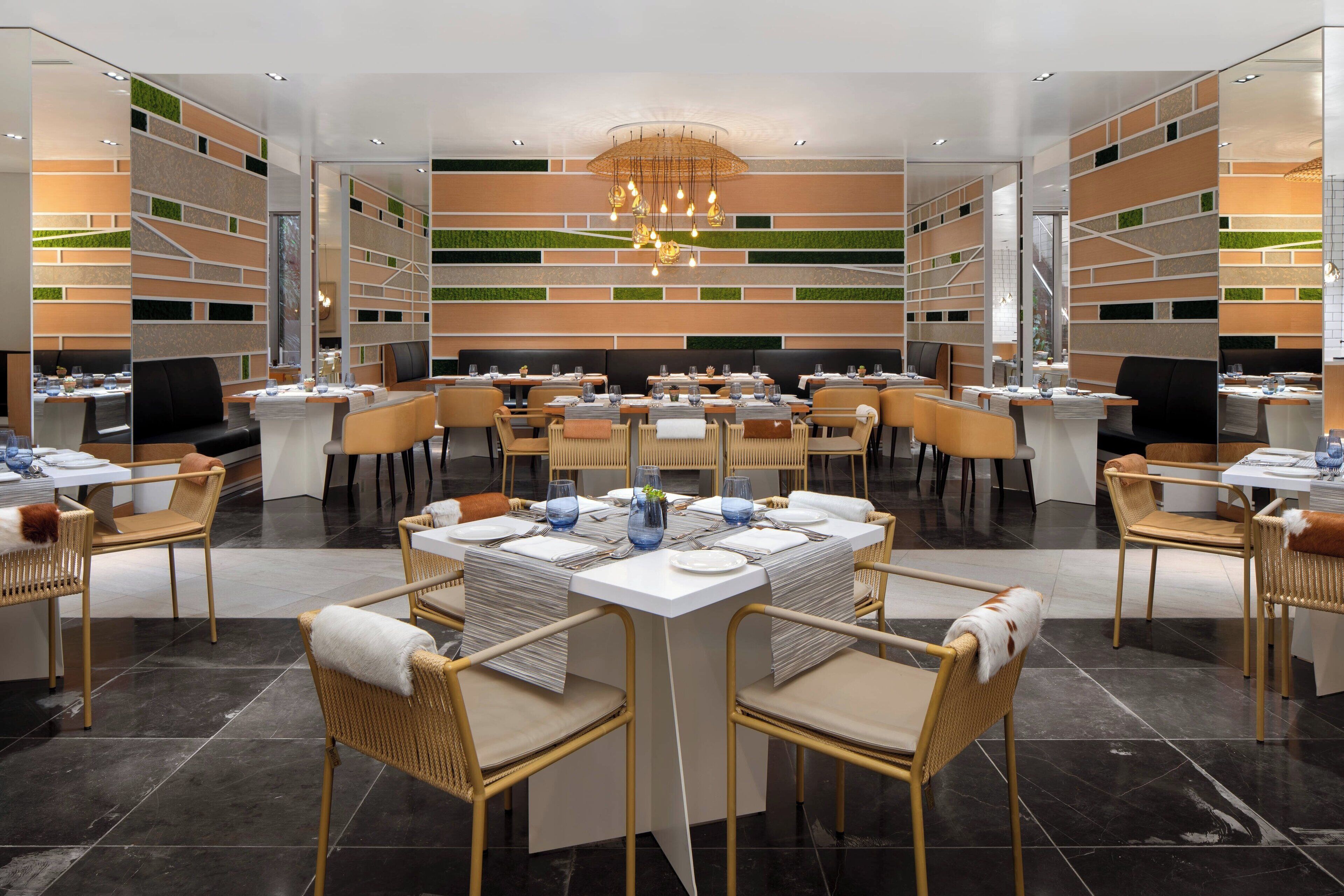 A modern dining area with multiple tables and chairs, a central chandelier, and a patterned wall design.