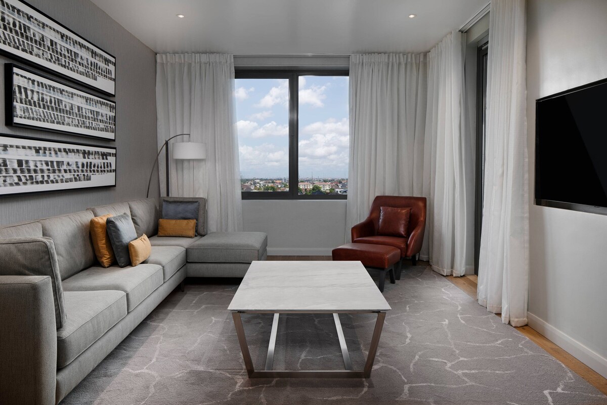 A modern living room with a grey sectional sofa, a red armchair, a white coffee table, and a large window offering a city view.
