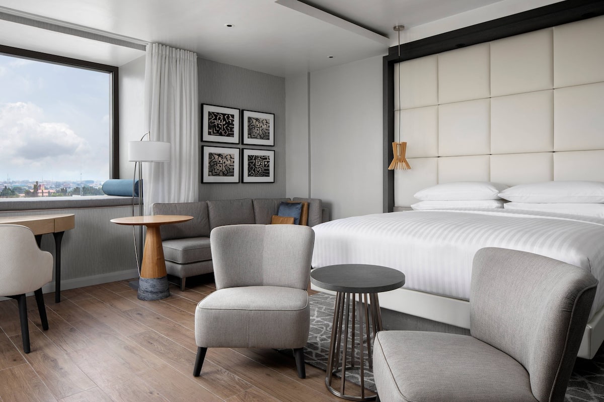 A modern hotel room with a large bed, a sofa, a small table, and a view of the city.