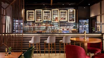 Un espace bar au design moderne, avec un comptoir où sont exposées différentes bouteilles et un présentoir réfrigéré. On y trouve des tabourets de bar et une table ronde avec une lampe.