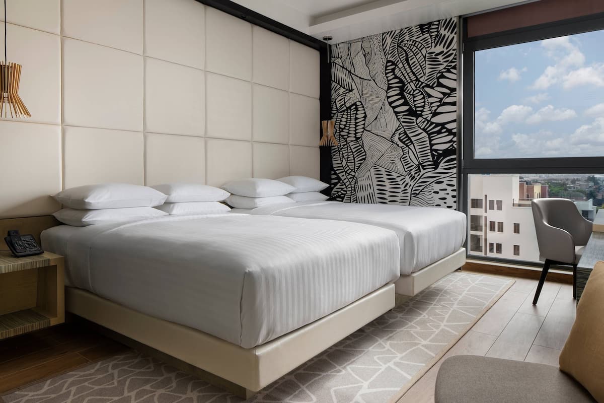 A modern hotel room with a large bed, a bedside phone, a patterned wall, and a view of the city through a large window.