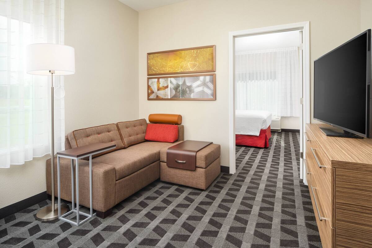 Ein Hotelzimmer mit einer Couch, einem Beistelltisch, einem Fernseher, einem Gemälde und einem gemusterten Teppich.