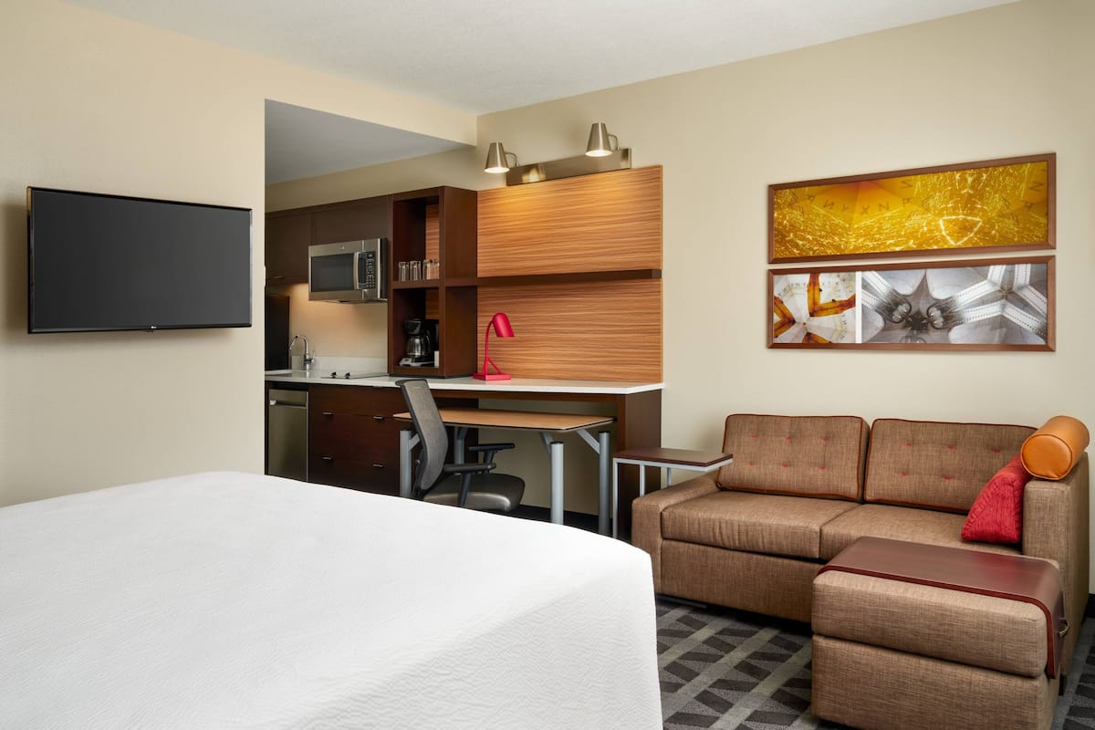 Ein Hotelzimmer mit Bett, Schreibtisch mit Stuhl, einer Küchenzeile, einer Couch und einem an der Wand befestigten Fernseher.