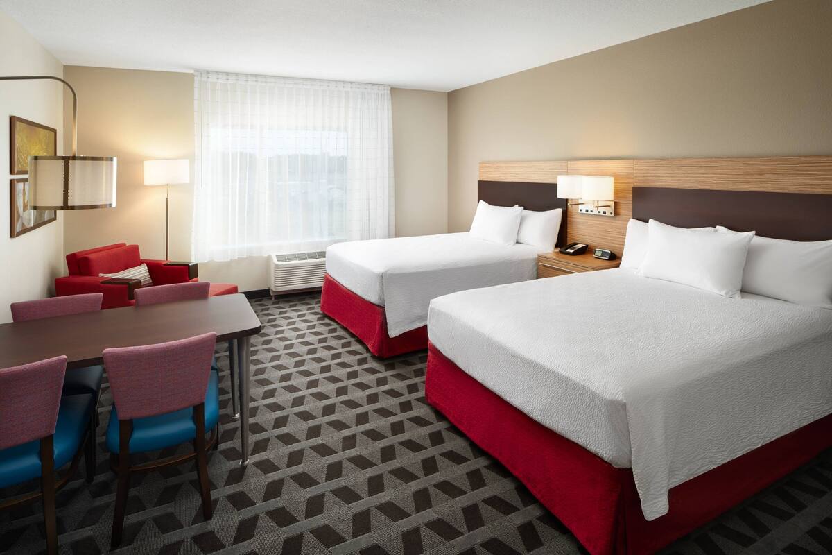 Ein Hotelzimmer mit zwei Betten, einem Esstisch mit Stühlen, einem roten Sofa und einem großen Fenster mit transparenten Vorhängen.