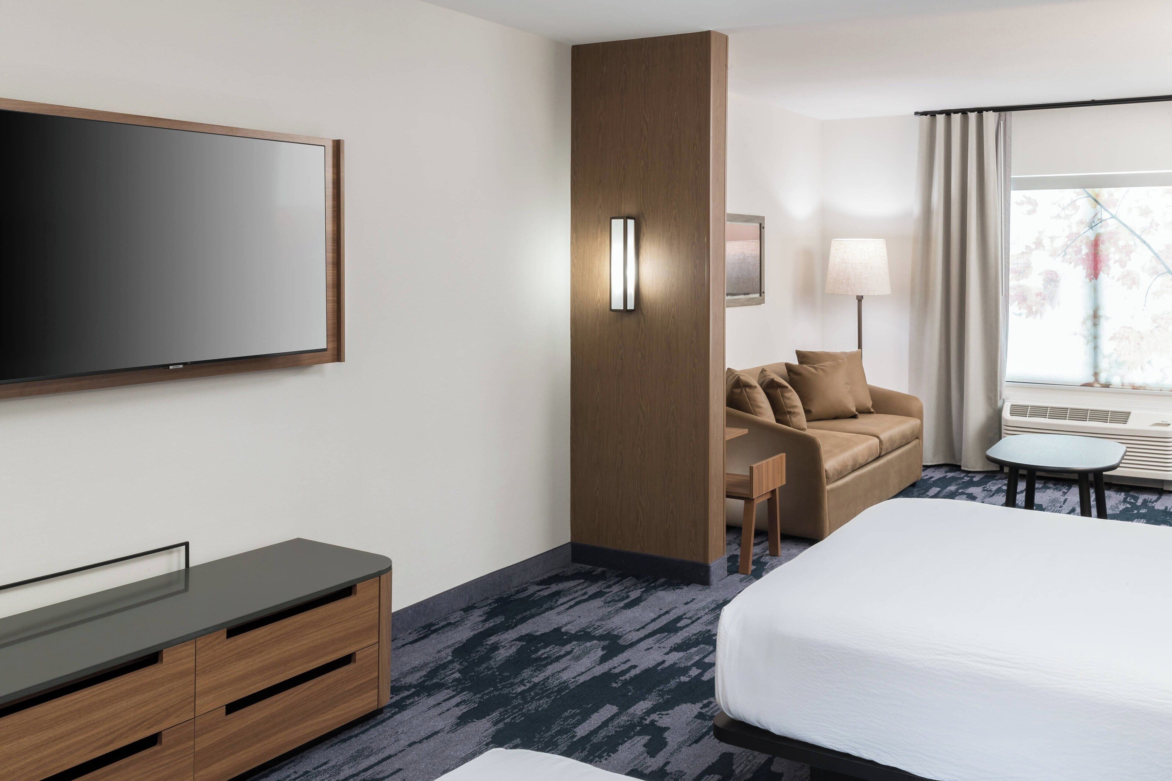 A modern hotel room with a large bed, a flat-screen TV, a wooden cabinet, a sofa, a small table, and a window with curtains.