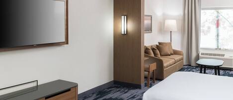 A modern hotel room with a large bed, a flat-screen TV, a wooden cabinet, a sofa, a small table, and a window with curtains.