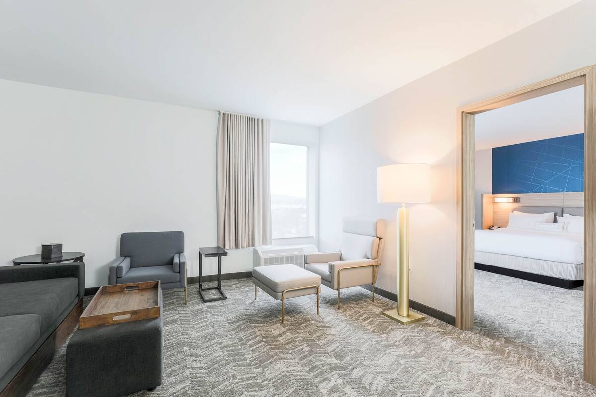 A hotel room with a grey sofa, two armchairs, a wooden coffee table, a floor lamp, and a bed with white bedding.