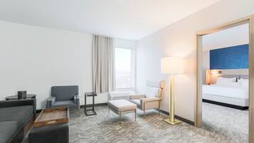 Quarto de hotel com sofá cinzento, duas poltronas, mesa de centro de madeira, abajur e cama com roupa de cama branca.