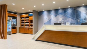 A reception area with a modern design, including a reception desk, a counter, and a wall with geometric patterns. There are wooden cabinets with various items and a refrigerator in the background. - SpringHill Suites by Marriott San Jose Fremont (Fremont)