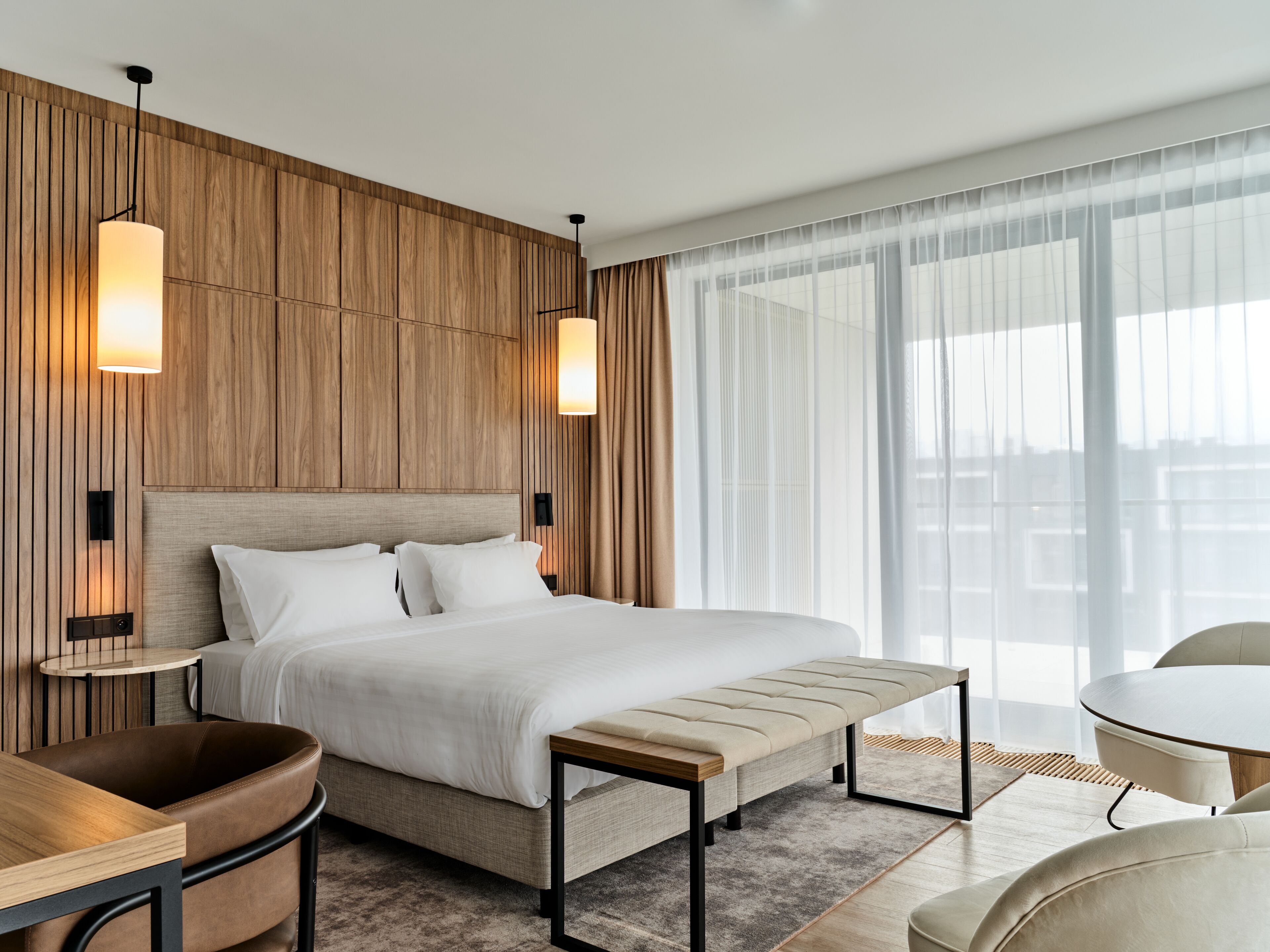 Habitación de hotel moderna con una cama grande, panel de pared de madera, dos lámparas colgantes, una mesa redonda y vistas de la ciudad a través de cortinas translúcidas.