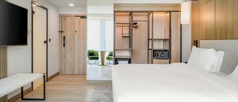 A modern hotel room with a large bed, a TV mounted on the wall, a built-in wardrobe, and a seating area with chairs.