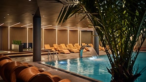A modern indoor pool area with lounge chairs, a water feature, and a potted plant.