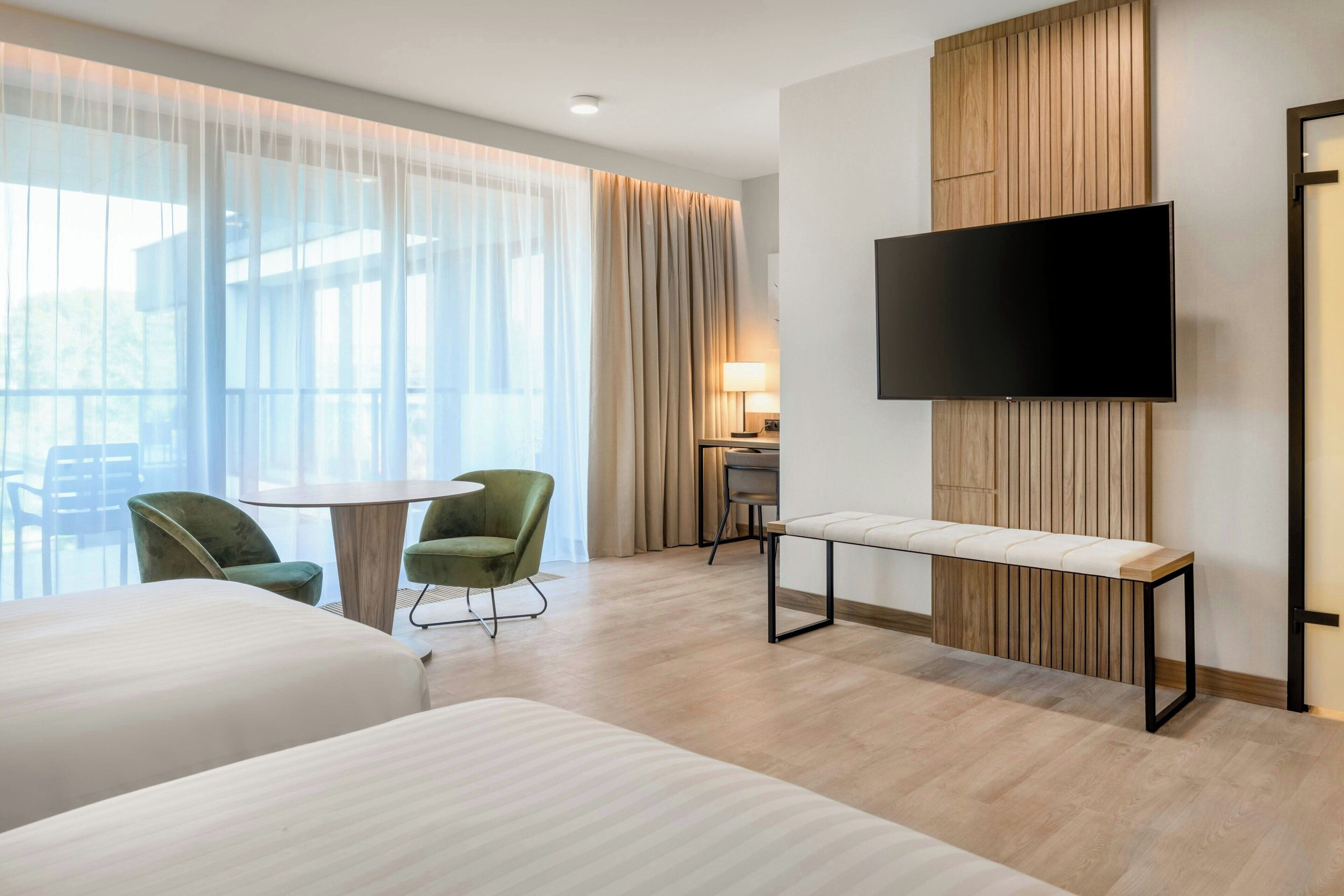 Camera d'albergo con un letto grande, un tavolino, due sedie verdi, una TV a parete e vista sull'esterno tramite tende trasparenti.