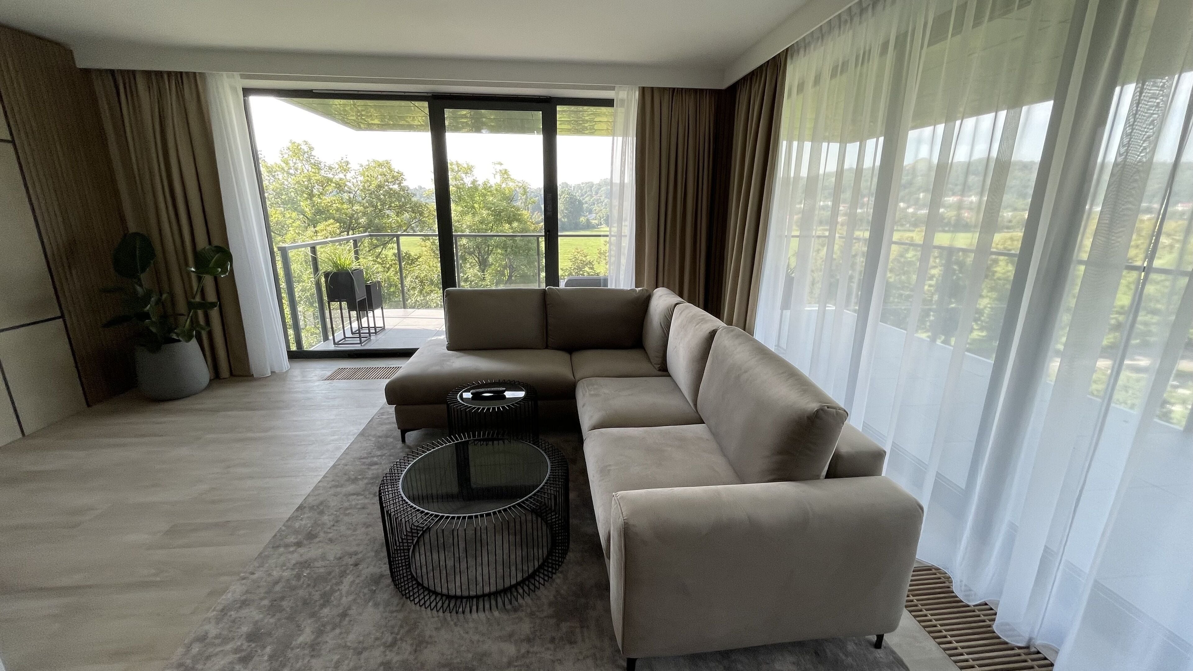 A modern living room with a sectional sofa, a round coffee table, and a balcony with a view of greenery.
