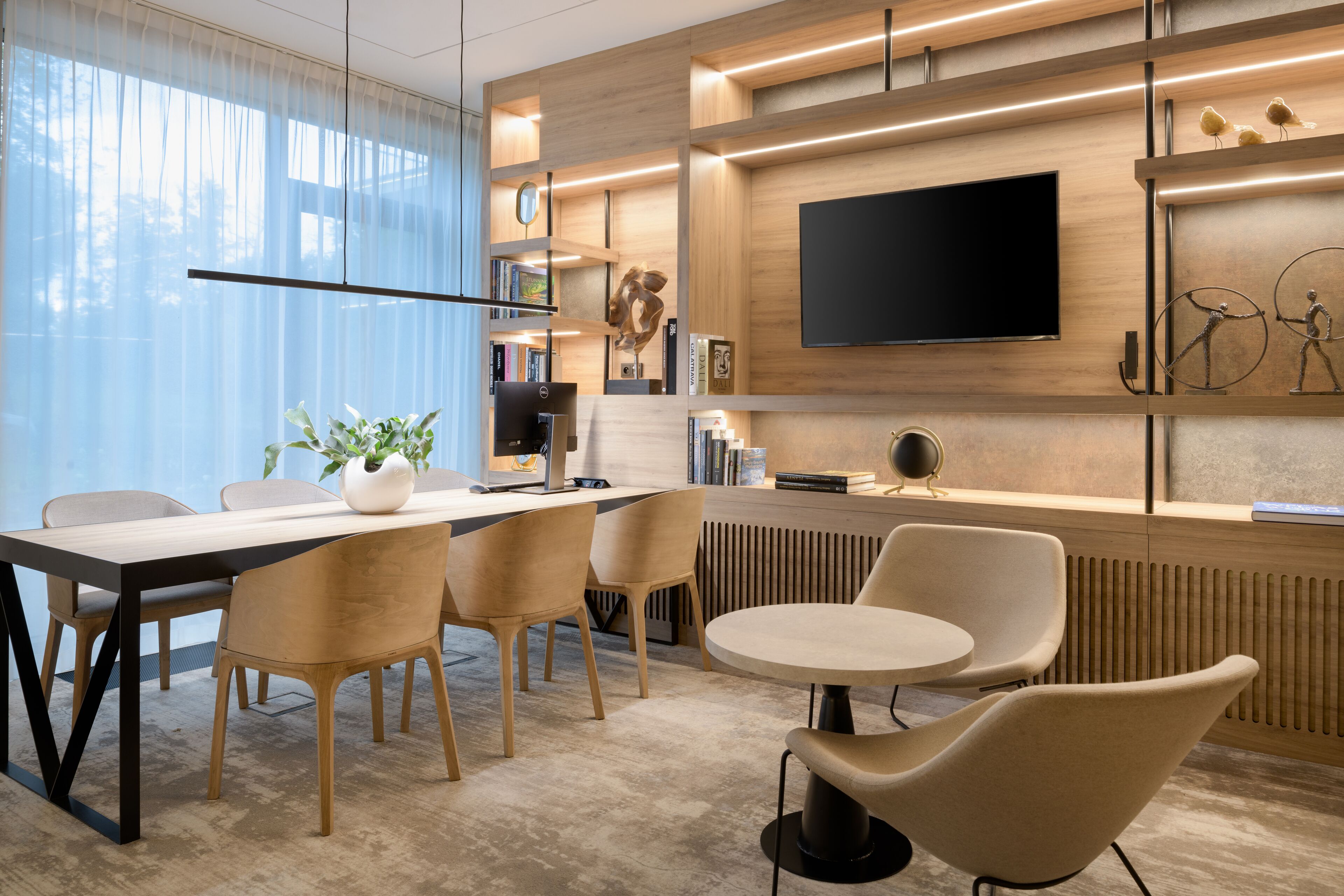 A modern office space with a long table, several chairs, a round table, and a TV mounted on the wall.