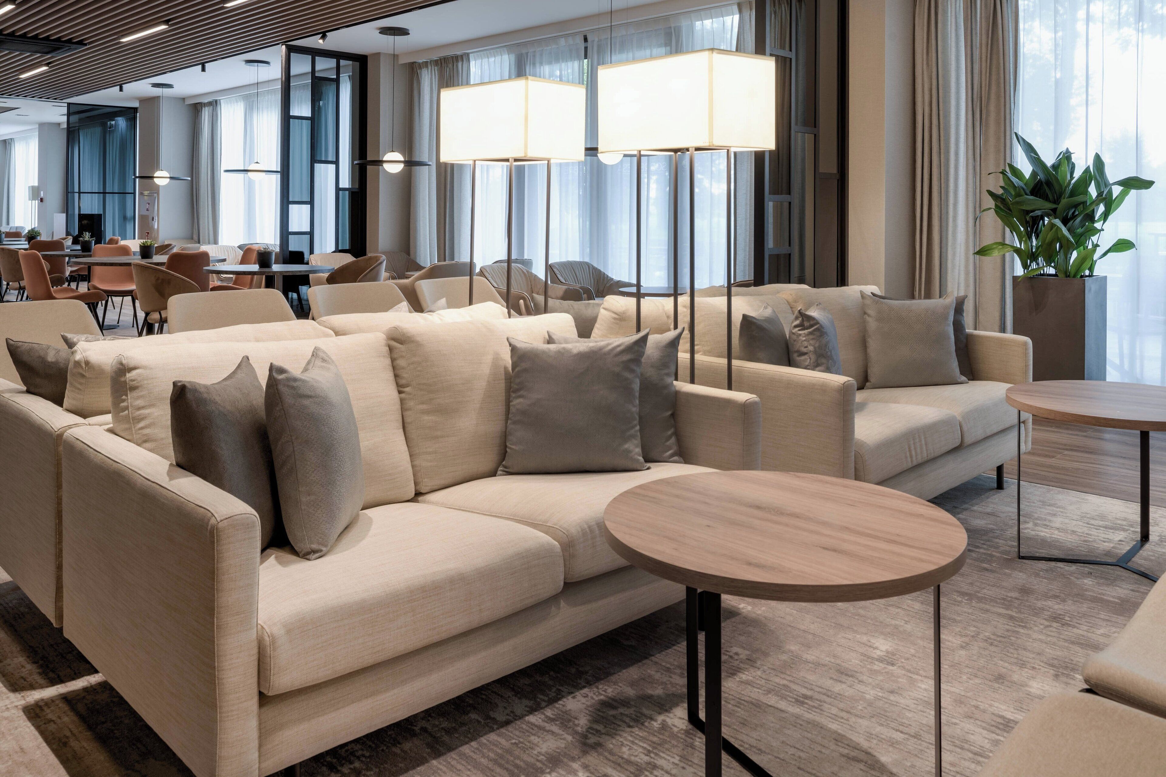 A modern hotel lobby with a beige sofa set, round wooden tables, and large floor lamps.