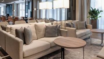 A modern hotel lobby with a beige sofa set, round wooden tables, and large floor lamps.
