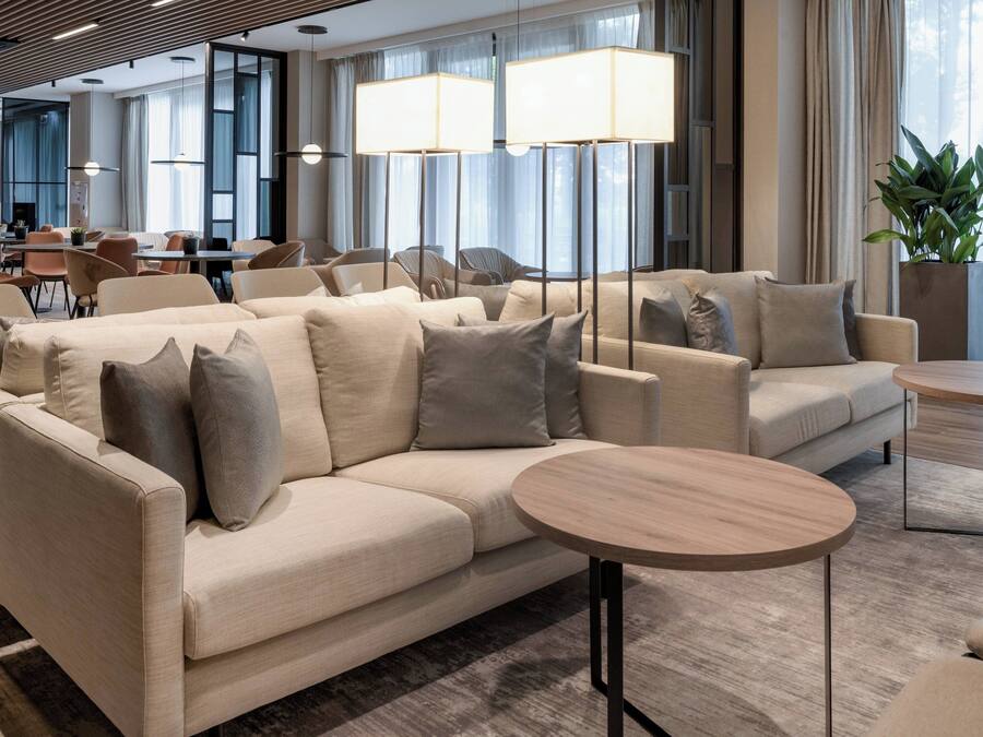A modern hotel lobby with a beige sofa set, round wooden tables, and large floor lamps.