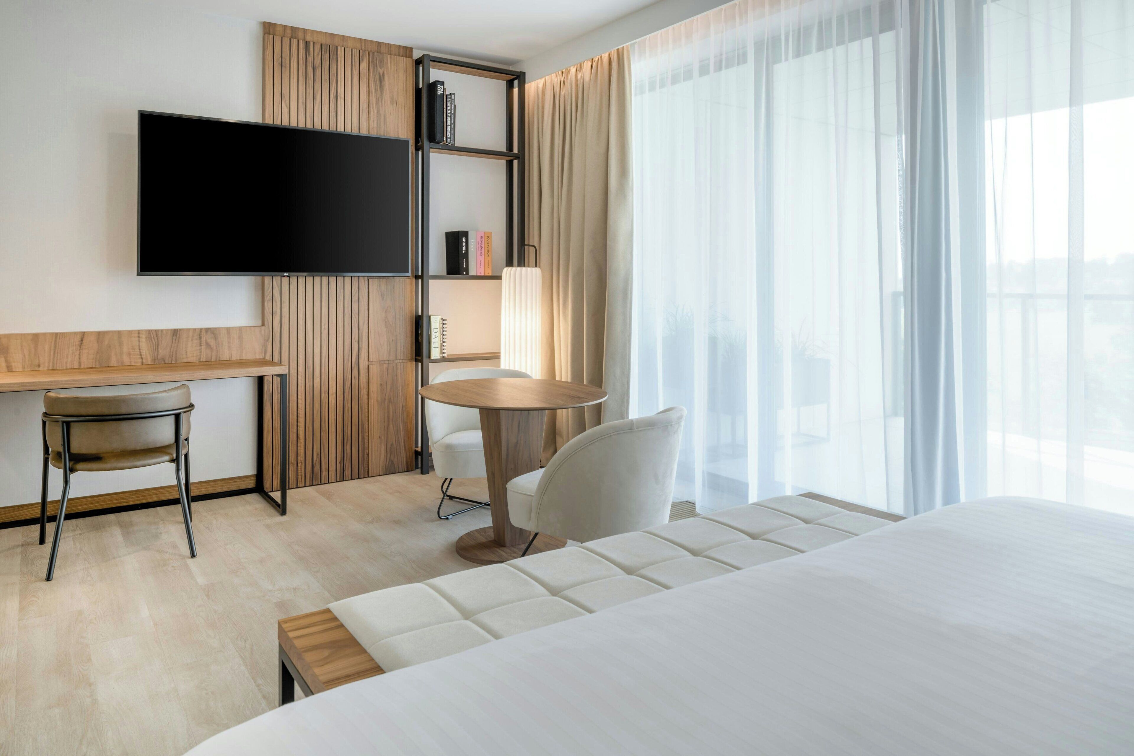 Quarto de hotel moderno com uma cama grande, uma escrivaninha com cadeira, uma mesa redonda, televisão fixada na parede e uma estante de livros.