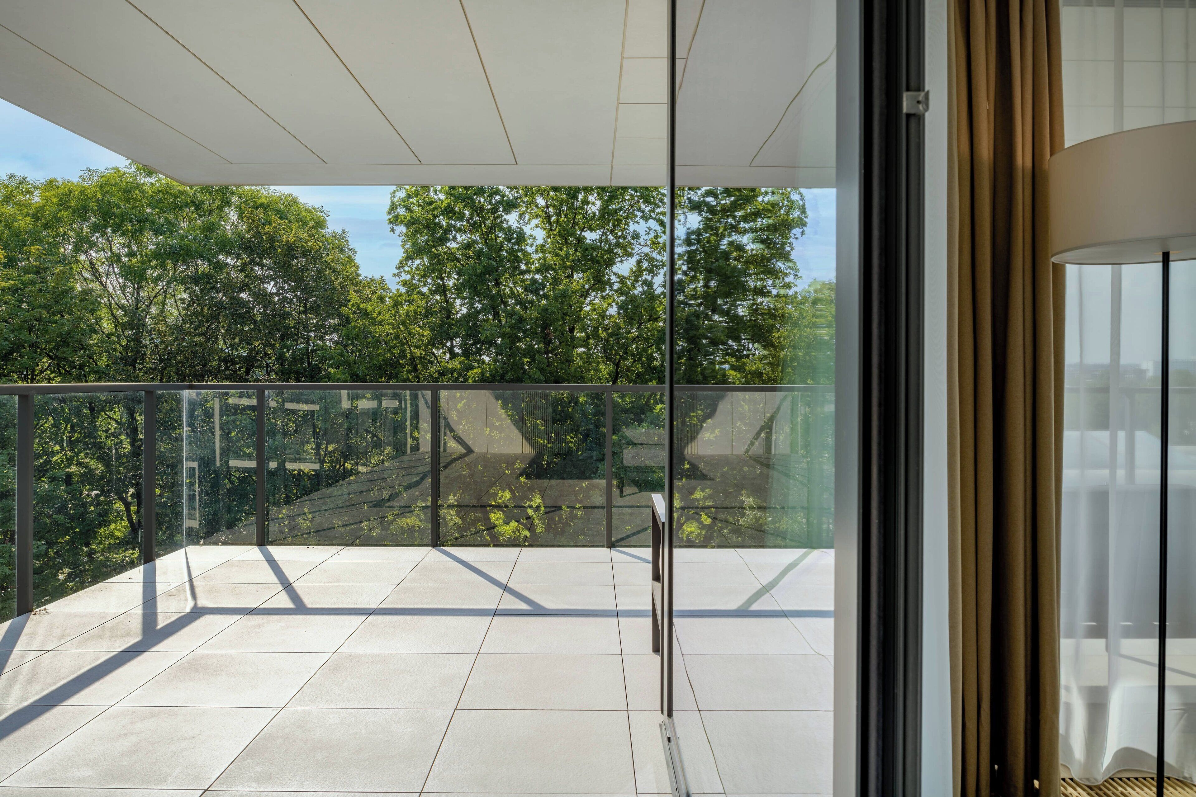 A modern balcony with glass railings, a tiled floor, and a view of trees and buildings.