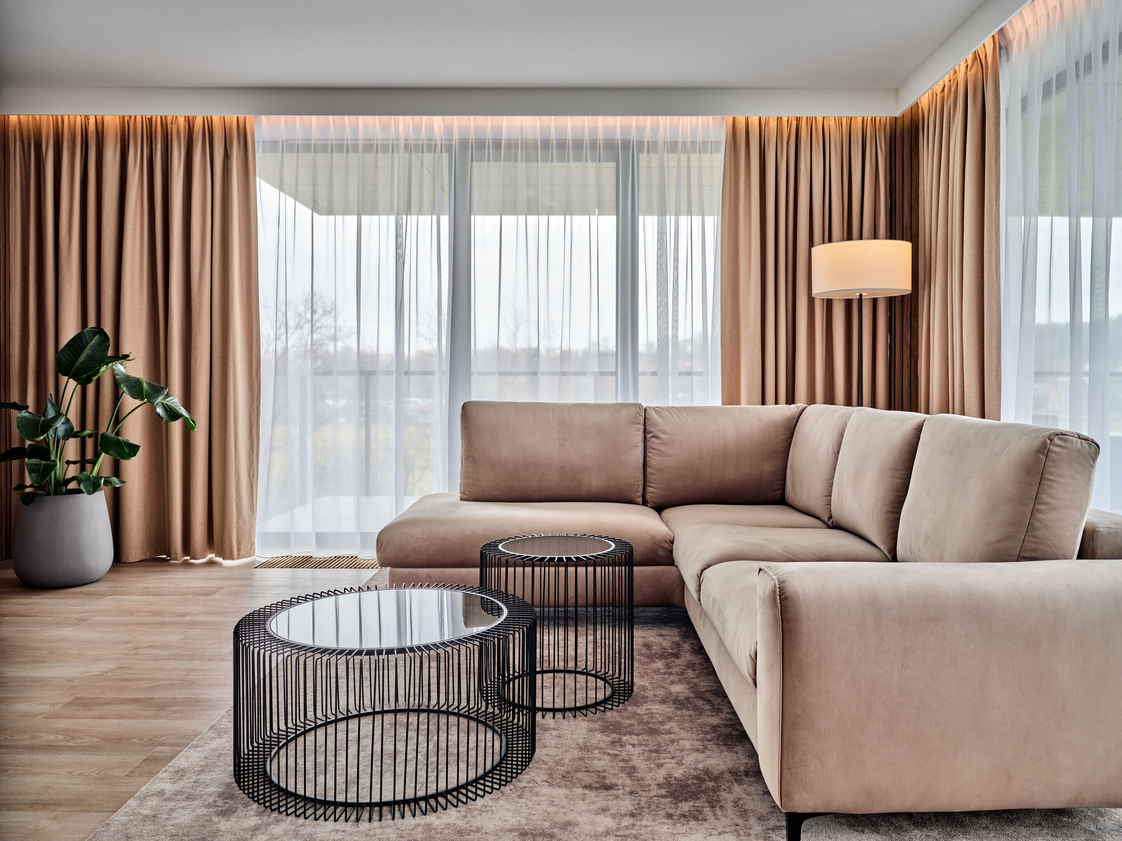A modern living room with a beige sectional sofa, two black wire coffee tables, a large potted plant, and a view of the city through large windows with sheer curtains.