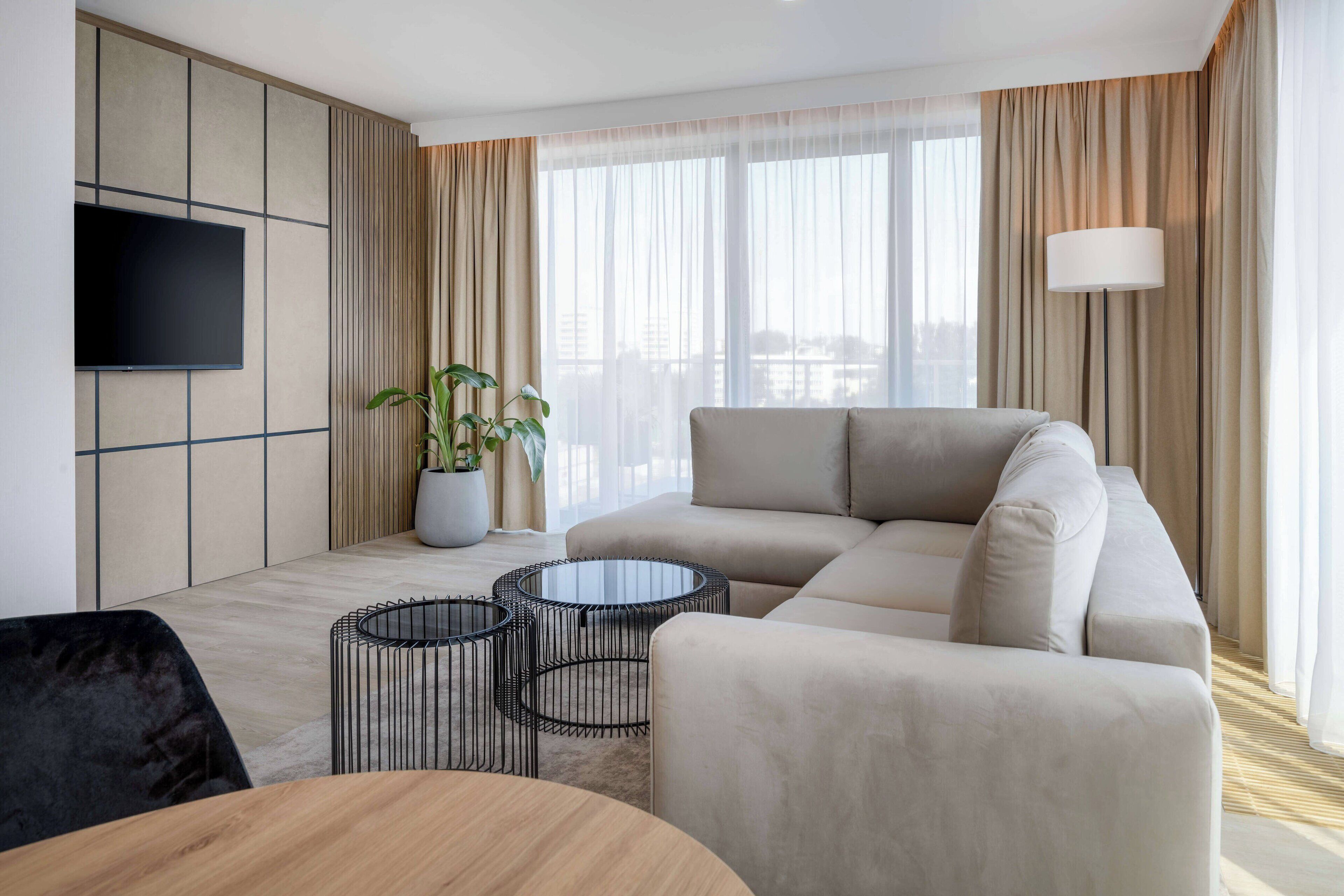 Un salon moderne avec un canapé d’angle, un téléviseur à écran plat fixé au mur, une plante en pot et une lampe sur pied.