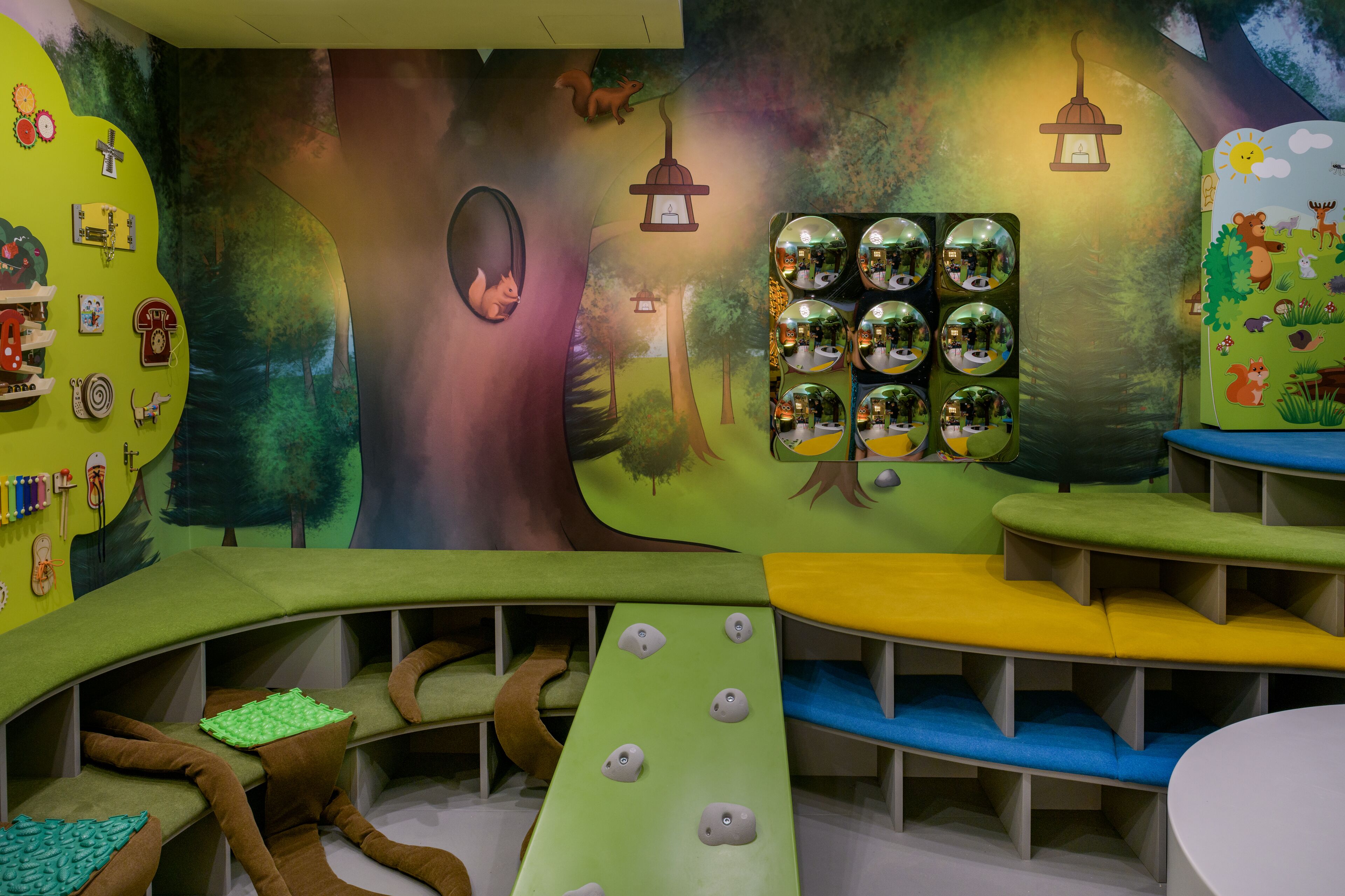 Zona infantil con asientos de colores y un mural que representa un paisaje forestal.