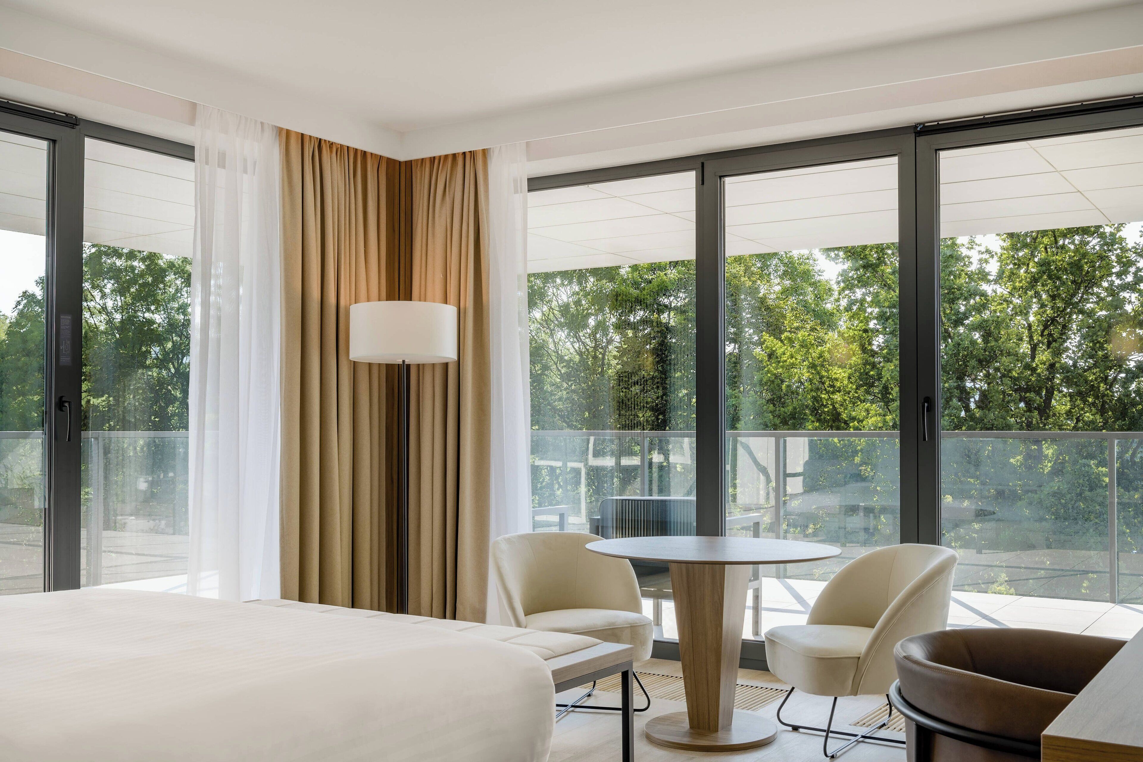 A hotel room with a large bed, two chairs, a small table, and a view of trees through large windows.