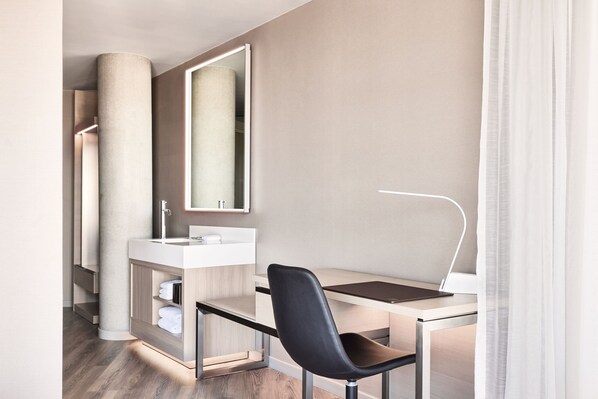 A modern hotel room with a minimalist design, featuring a desk, a chair, a large mirror, and a built-in vanity with a sink.