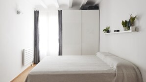 Een minimalistische slaapkamer met een bed, een radiator, gordijnen, een kledingkast en een plank met planten en een klok.
