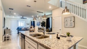 A spacious kitchen with granite countertops, stainless steel sinks, and pendant lighting. A dining area with a blue sofa and a staircase leading to an upper level is visible in the background. - WIR Free Pool Heat/Grill Use, Walk to Lazy River (Four Corners)