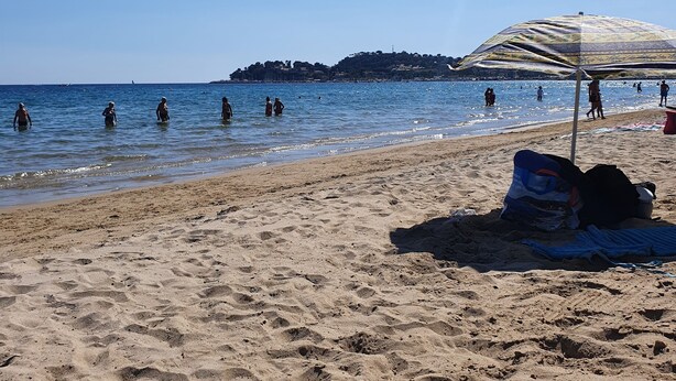 Ein Strand mit badenden und spazierenden Personen, einem großen Sonnenschirm und einer entfernten Insel.