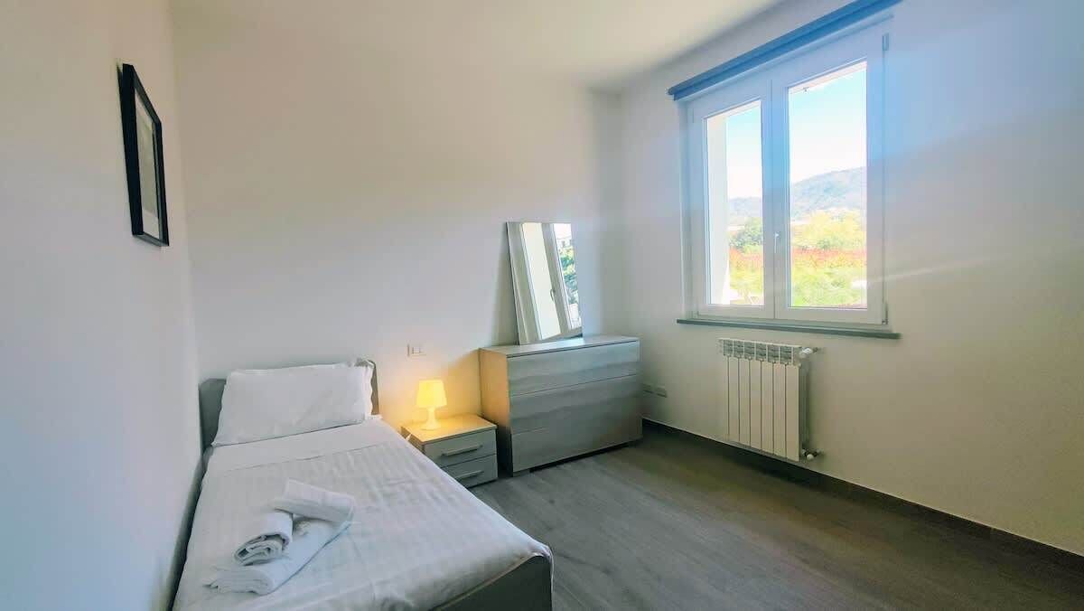 Camera da letto con un letto, un comodino, una lampada, una cassettiera e una finestra vista su spazi verdi.
