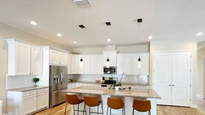 Una cocina moderna con gabinetes blancos, una isla central con taburetes altos, electrodomésticos de acero inoxidable e iluminación colgante.