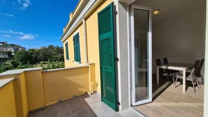 A balcony with a yellow exterior, green shutters, and a glass door leading to an outdoor dining area with a table and chairs. - Balcony Condo Steps to Beach & Train Station - O (Levanto)