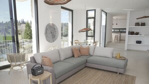 Ein modernes Wohnzimmer mit einem grauen Ecksofa, einem kleinen runden Tisch und einem Korbstuhl. Ein großes Fenster bietet einen Blick ins Freie.