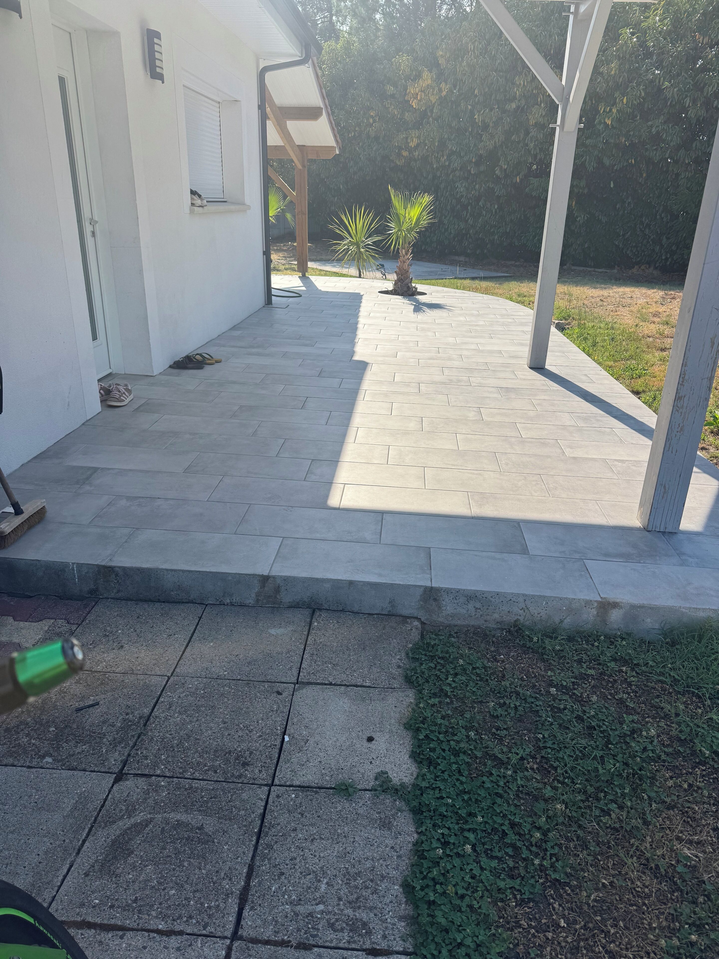 Une terrasse dallée avec un mur blanc, une porte et une fenêtre. On y trouve deux plantes en pot et un balai.