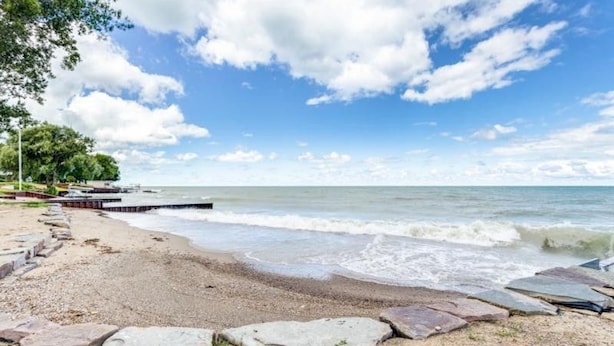 Ein Strand mit einem hölzernen Steg, der sich in ruhiges Wasser erstreckt, umgeben von felsiger Küste und Bäumen.